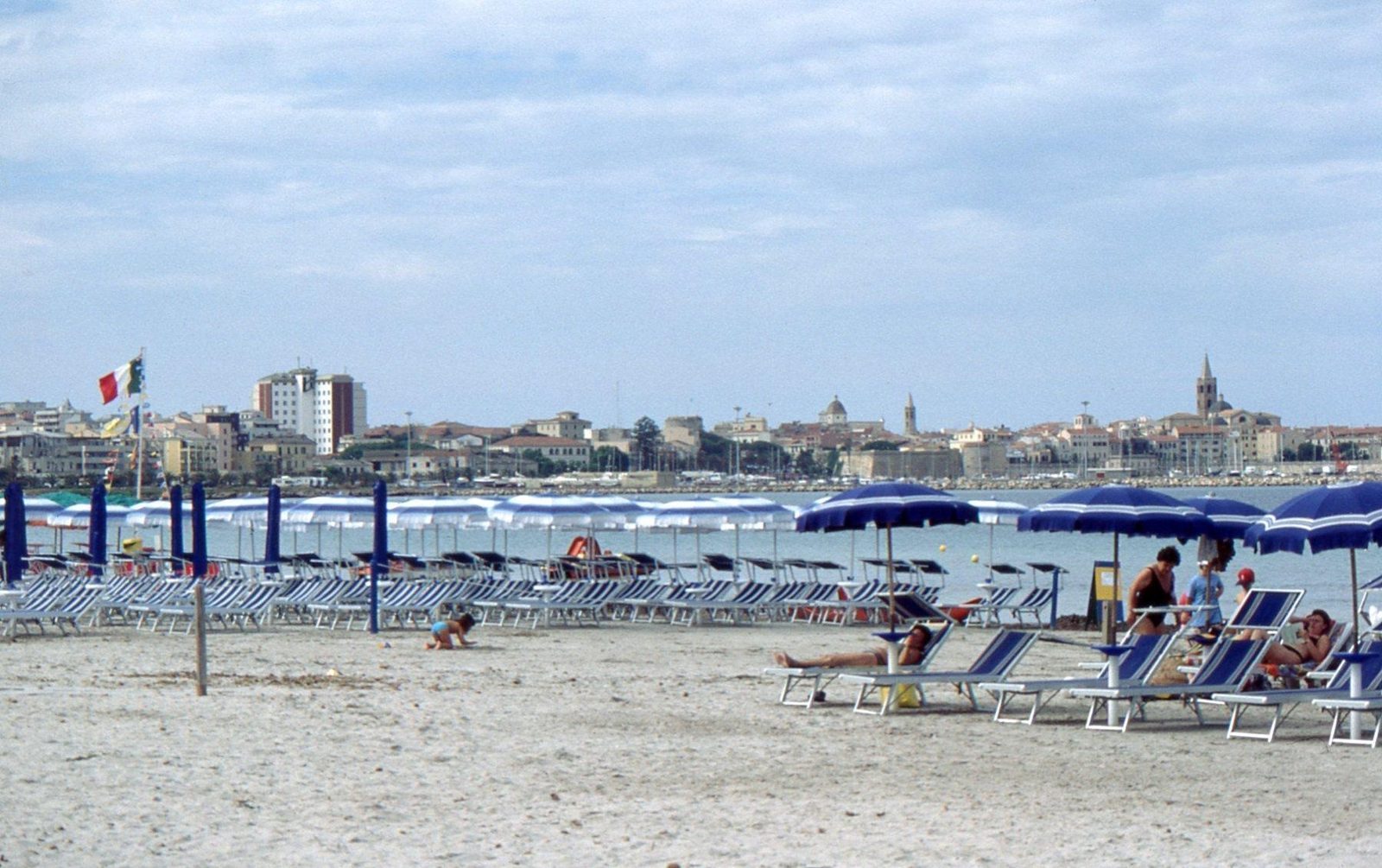 Spiaggia attrezzata con ombrelloni blu sul lungomare di Alghero, con il profilo della città sullo sfondo
